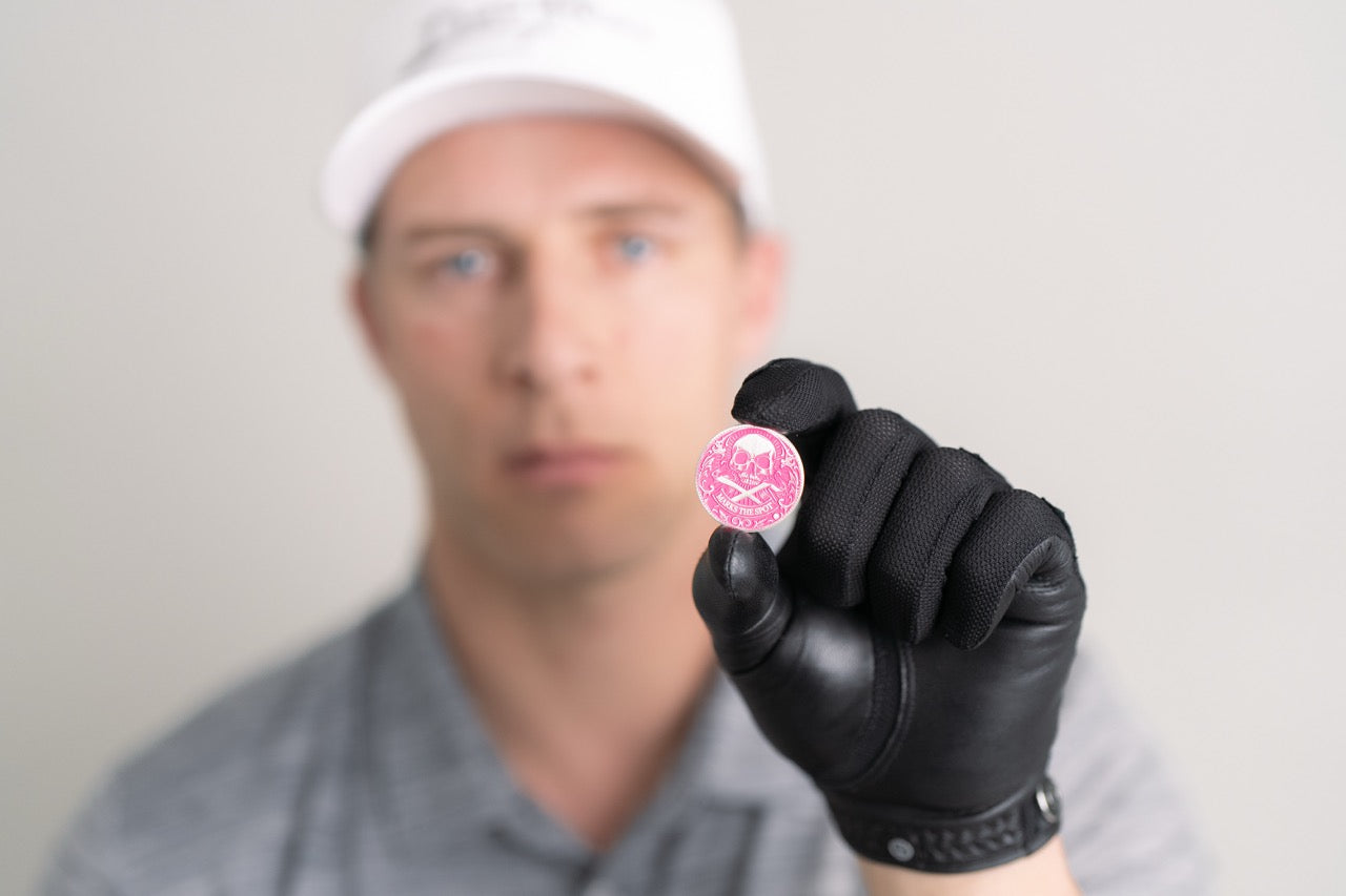 A person in a white cap and gray shirt, wearing a black glove, holds up Doubloon's The Silver Omen Antique, Color Edition golf ball marker—a pink coin with graphic design—while their face is out of focus in the background.