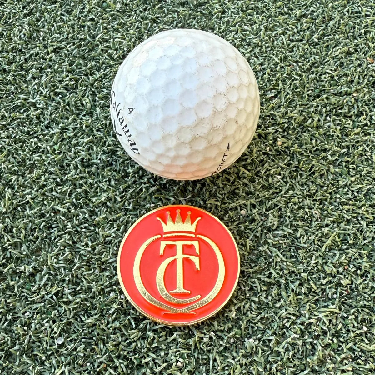 A white golf ball sits on artificial grass beside the Grunk Dolfer Tour Champ Ball Marker—red, circular, with a gold crown and T/C design—ideal for magnetic hat clips and thoughtful golf gifts.