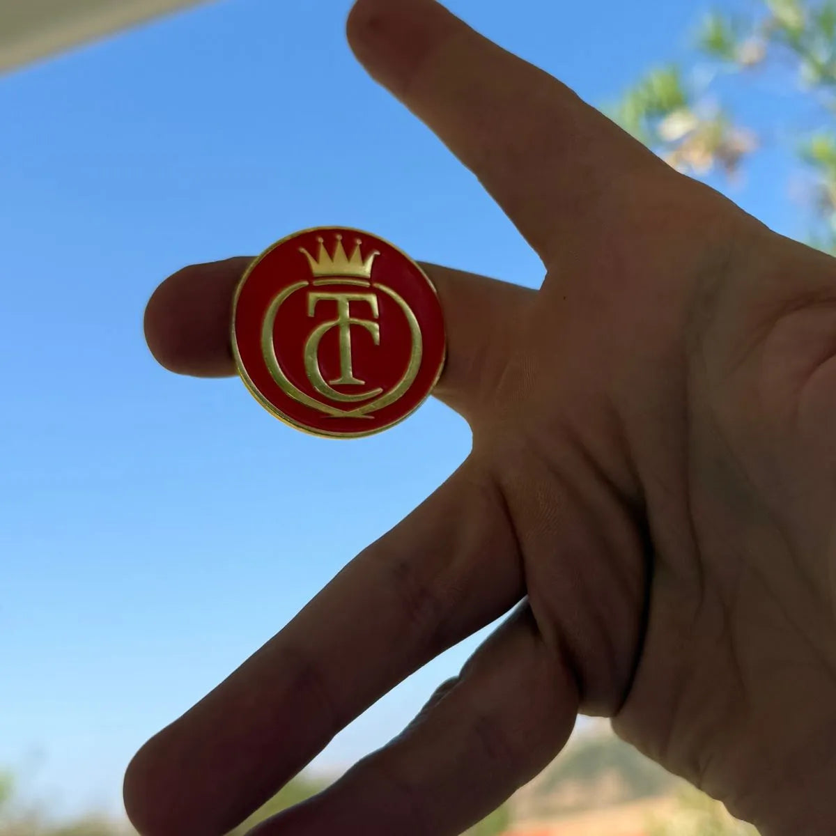A hand holds the Grunk Dolfer Tour Champ Ball Marker, a round red and gold pin with a crown and T/C logo—ideal as a magnetic hat clip or unique golf gift—set against a blue sky and green landscape.